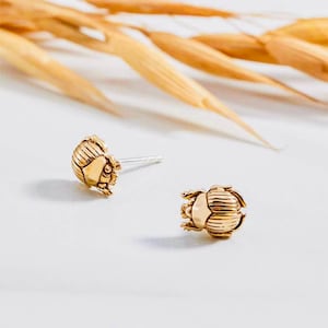 May include: A pair of gold-toned earrings featuring a detailed scarab beetle design. The earrings are small and delicate, perfect for adding a touch of whimsy to any outfit.