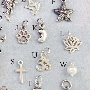 May include: A collection of silver charms, including a fish, a bird, a starfish, a paw print, a crescent moon, a lotus flower, a cross, an Om symbol, a pearl, a Celtic knot, and a bell.