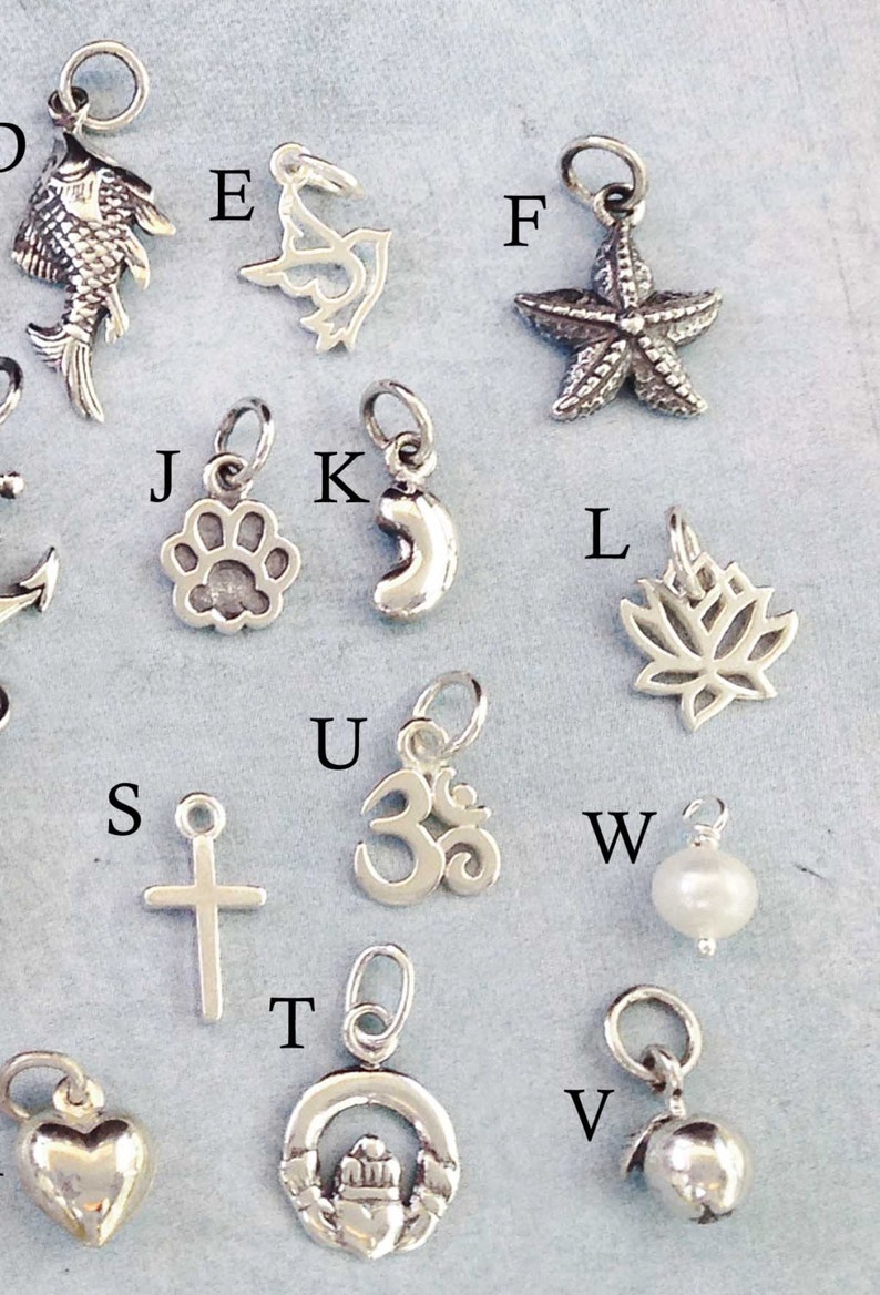May include: A collection of silver charms, including a fish, a bird, a starfish, a paw print, a crescent moon, a lotus flower, a cross, an Om symbol, a pearl, a Celtic knot, and a bell.