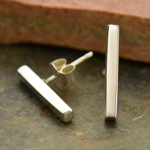 May include: A pair of silver bar stud earrings. Each earring is a rectangular bar with rounded edges and a post for piercing. The earrings are displayed on a textured, dark surface with a blurred background.