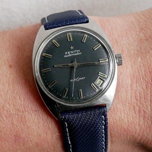 May include: A vintage Zenith AutoSport watch with a dark gray face and a navy blue fabric band. The watch has a silver-colored case and a date display at the 3 o'clock position. The watch band is approximately 0.75 inches wide.