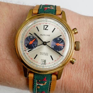 May include: A vintage gold-toned AURORE chronograph watch with a silver face, black markers, and two sub-dials. The watch features a green and tan fabric band with a floral pattern. The date is set to 22.