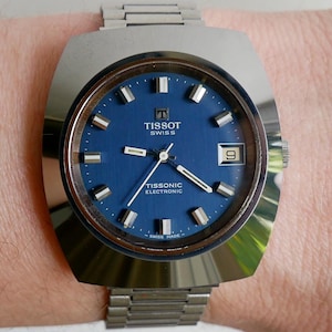 Puede incluir: Un reloj Tissot Tissonic Electronic plateado con una esfera azul y agujas plateadas. El reloj tiene una caja cuadrada y una correa de metal plateado.