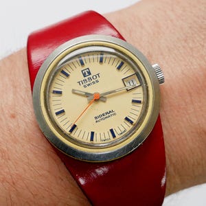 Könnte beinhalten: Eine Vintage Tissot Sideral Automatik-Armbanduhr mit cremefarbenem Zifferblatt, blauen Markierungen und Datumsfenster. Die Uhr hat ein silberfarbenes Gehäuse und ein leuchtend rotes Lederarmband. Auf dem Zifferblatt ist das Tissot Swiss Logo zu sehen.