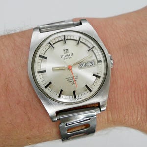Könnte beinhalten: Eine silberfarbene Tissot Seastar PR 516 GL Automatik-Uhr. Das Zifferblatt ist silberfarben mit schwarzen Stundenmarkierungen, Datumsanzeige und rotem Sekundenzeiger. Die Uhr hat ein silberfarbenes Metallarmband.