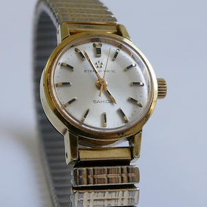 May include: A gold-toned wristwatch with a white face and black markings. The watch face says "ETERNA-MATIC" and "SAHIDA". The watch has a gold-toned metal band.
