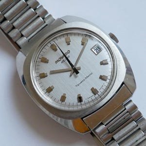 Pode incluir: Um relógio de pulso Movado Tempo-Matic prateado com um mostrador branco, ponteiros pretos e uma janela de data que mostra o número 28. O relógio tem uma pulseira de metal prateado.