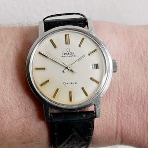 May include: A vintage Omega Genève automatic wristwatch with a silver-tone case and a black leather strap. The watch face is cream-colored with gold-tone hour markers and hands. The watch displays the Omega logo and the word "Genève".