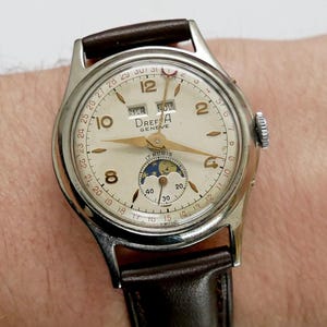 May include: A vintage Drefa Geneva wristwatch with a cream-colored face, gold hands, and a brown leather strap. The watch features a moon phase indicator and day/date display. The watch face displays the text "Drefa Geneva" and "17 Rubis".