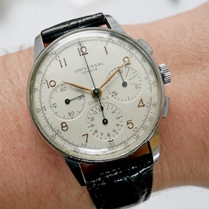 Peut inclure: Une montre chronographe Universal Geneve Compax vintage avec un cadran crème, un bracelet en cuir noir et un boîtier argenté. La montre comporte plusieurs sous-cadrans et des aiguilles dorées. Un accessoire élégant.
