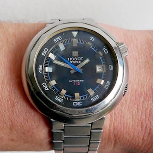 Puede incluir: Un reloj automático suizo Tissot vintage con correa y caja de acero inoxidable. La esfera del reloj es azul oscuro con detalles blancos y azules, incluyendo el logotipo de Tissot y el texto "AUTOMATIC T12". El reloj está en una muñeca.