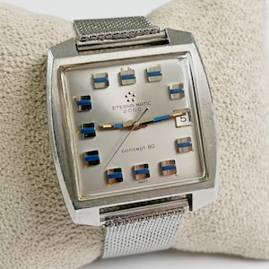 Puede incluir: Un reloj Eterna-Matic 2000 Concept 80 plateado con esfera rectangular. El reloj presenta una esfera plateada con detalles azules y plateados, visualización de fecha y correa de malla. Fabricado en Suiza.