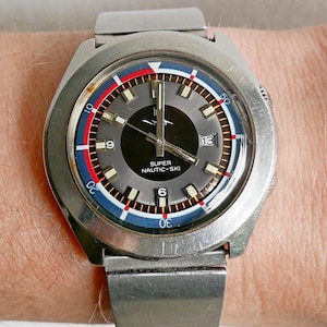 Puede incluir: Un reloj vintage plateado con esfera gris, con detalles en rojo, blanco y azul. El reloj tiene una pantalla de fecha a las 3 en punto y el texto "SUPER NAUTIC-SKI". La correa del reloj también es plateada.