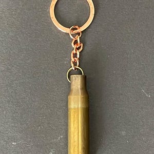 Puede incluir: Un llavero de latón con forma de casquillo de bala con un anillo y una cadena de color cobre. El casquillo de la bala es de color marrón dorado y tiene un brillo metálico. El llavero está diseñado para sujetarse a llaves o bolsos.