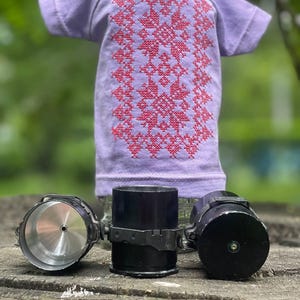 Op de afbeelding: Een lavendelkleurig T-shirt met een rood geborduurd ontwerp, weergegeven op een heldere fles. Daaronder zijn drie zwarte cilindrische objecten met zilveren en zwarte details gerangschikt op een houten oppervlak. De achtergrond is vaag groen.