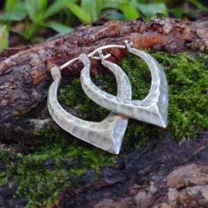 May include: Silver-coloured hoop earrings with a hammered texture. The earrings have a distinctive, pointed shape and are displayed on a mossy, wooden surface. The background features green foliage.