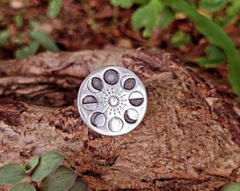 May include: A silver-colored ring featuring a celestial design. The ring's face showcases a sun in the center, surrounded by phases of the moon. The ring is set against a natural wood background with green foliage.