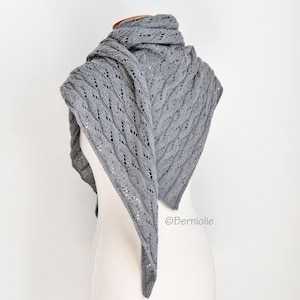 Puede incluir: Un chal triangular gris con un delicado diseño calado. El chal está drapeado sobre un maniquí, mostrando su intrincado patrón de punto y su suave textura. El color del chal es un gris apagado.