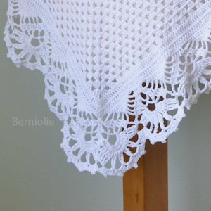 May include: Close-up of a white, crocheted textile with a delicate floral lace border. The intricate openwork design creates a textured pattern. The text "Berniolie" is visible in the lower left corner.