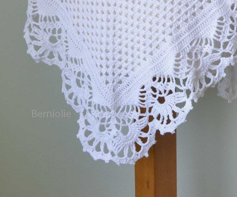 May include: Close-up of a white, crocheted textile with a delicate floral lace border. The intricate openwork design creates a textured pattern. The text "Berniolie" is visible in the lower left corner.