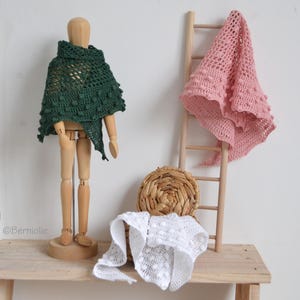 Puede incluir: Un maniquí de madera exhibe un chal de ganchillo verde oscuro con detalles de bolitas. Un chal rosa de ganchillo cuelga de una pequeña escalera de madera. También se ven un chal blanco de ganchillo y una cesta de mimbre. Los chales están hechos a mano.
