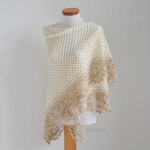May include: Cream-colored crocheted shawl with a decorative border in a light tan color. The shawl is draped over a mannequin, showcasing the intricate openwork design and delicate lace trim. The shawl is a light and airy accessory.