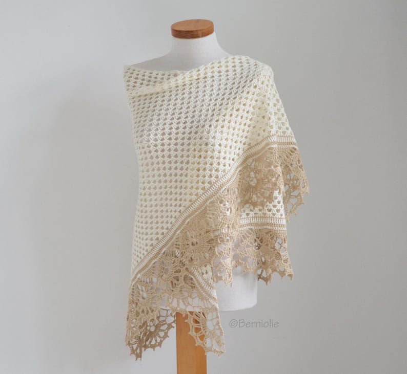 May include: Cream-colored crocheted shawl with a decorative border in a light tan color. The shawl is draped over a mannequin, showcasing the intricate openwork design and delicate lace trim. The shawl is a light and airy accessory.