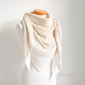 May include: Cream-colored knitted shawl draped over a mannequin. The shawl features a textured pattern and a triangular shape, with the ends hanging down. The color is a soft off-white, suitable for various outfits. The shawl is displayed on a white background.