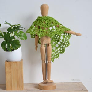 Puede incluir: Un chal de ganchillo verde lima, con un delicado diseño calado, está drapeado sobre un maniquí de artista de madera. El chal tiene un borde festoneado. Una planta en maceta en una maceta blanca está a la izquierda del maniquí.