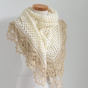 Puede incluir: Chal de crochet color crema con un borde festoneado en color beige. El chal está drapeado sobre un maniquí, mostrando su diseño calado y sus intrincados detalles. La textura y el color del chal sugieren un accesorio delicado y hecho a mano.