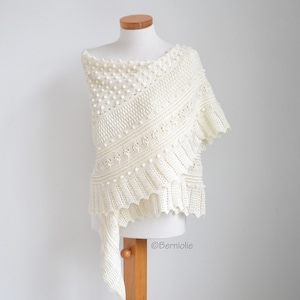 May include: An off-white crochet shawl featuring a textured pattern of small raised bobbles, diagonal ribbed sections, and a scalloped edge. The garment is displayed on a mannequin and includes the text Berniolie printed on the lower right side.