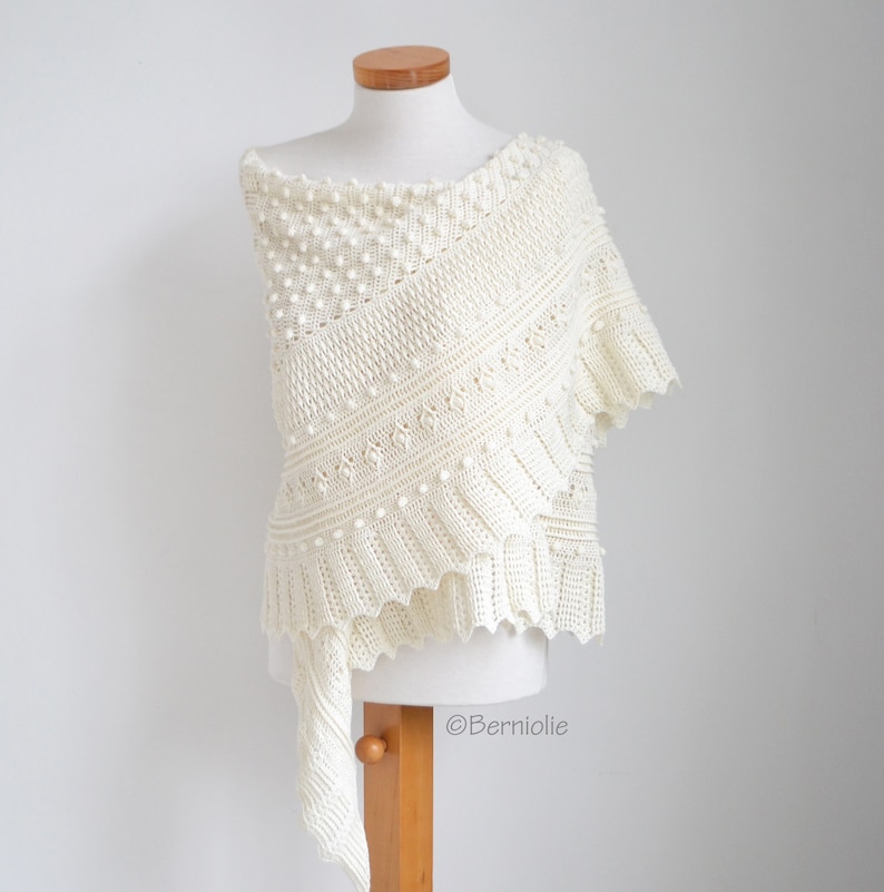 May include: An off-white crochet shawl featuring a textured pattern of small raised bobbles, diagonal ribbed sections, and a scalloped edge. The garment is displayed on a mannequin and includes the text Berniolie printed on the lower right side.