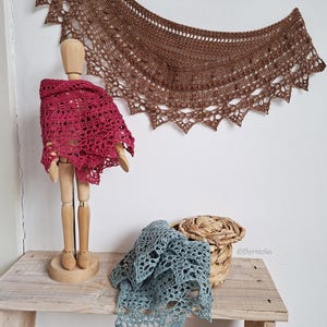 Puede incluir: Exhibición de chales de ganchillo hechos a mano en varios colores. Un chal rojo oscuro está drapeado sobre un maniquí de madera, mientras que un chal marrón cuelga de la pared. Un chal azul-verdoso y una cesta de mimbre están sobre una superficie de madera.