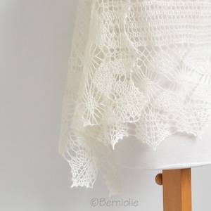 Puede incluir: Chal de color marfil, tejido abierto con un delicado patrón de encaje. El chal está drapeado sobre una forma blanca, mostrando sus intrincados diseños florales y geométricos. La tela parece suave y ligera, con un borde festoneado.