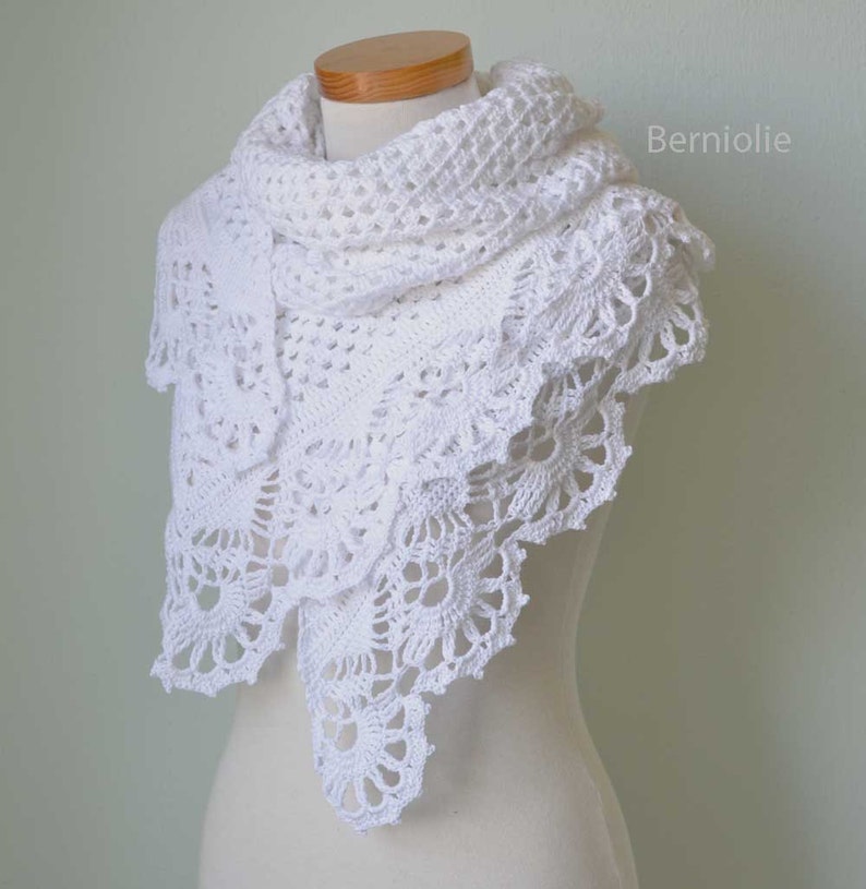 May include: A white, crocheted shawl with a delicate, lacy design. The shawl is draped over a mannequin, showcasing its intricate floral and geometric patterns. The edges feature a scalloped trim, adding a touch of elegance. The word "Berniolie" is visible on the right side.
