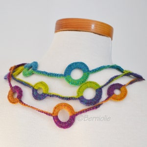 Op de afbeelding: Een veelkleurige gehaakte ketting met een reeks verbonden cirkels. De ketting is gemaakt met verschillende tinten blauw, groen, paars en oranje garen.