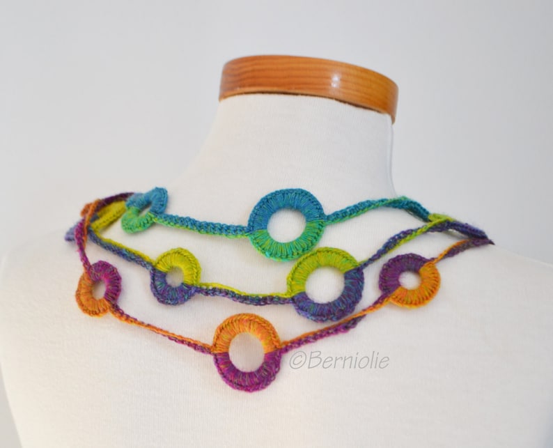 Op de afbeelding: Een veelkleurige gehaakte ketting met een reeks verbonden cirkels. De ketting is gemaakt met verschillende tinten blauw, groen, paars en oranje garen.