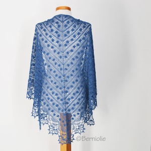 May include: A blue crocheted shawl displayed on a wooden mannequin. The shawl features a delicate openwork design with a geometric pattern of diamonds and circles. The edges are scalloped, adding a decorative touch. The shawl is a vibrant shade of blue.