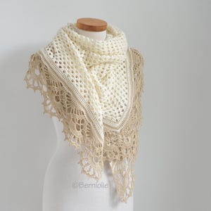 May include: A cream-colored, handmade crochet shawl with a delicate openwork design. The shawl features a scalloped, tan-colored border and is draped over a mannequin. The shawl is a triangular shape and appears soft and lightweight.