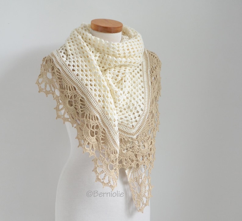 May include: A cream-colored, handmade crochet shawl with a delicate openwork design. The shawl features a scalloped, tan-colored border and is draped over a mannequin. The shawl is a triangular shape and appears soft and lightweight.