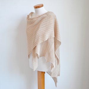 Puede incluir: Un chal tejido beige drapeado sobre un maniquí. El chal presenta un patrón texturizado y un diseño drapeado, con cuello de cisne y bordes puntiagudos. El color neutro y el tejido abierto sugieren un accesorio ligero y versátil.