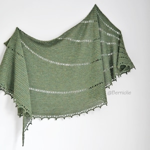 May include: A green crocheted shawl with a lacy border. The shawl is draped over a white wall. The text "@Berniolie" is visible in the lower right corner of the image.