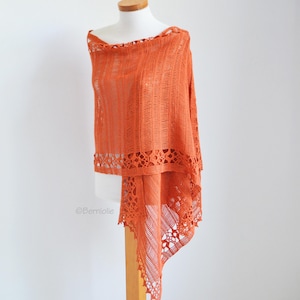 May include: An orange shawl displayed on a mannequin. The shawl is made of a lightweight, open-weave fabric with a textured pattern. The edges are finished with a decorative, openwork border. The shawl is draped to showcase its design and texture.
