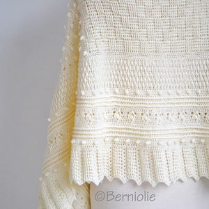 May include: A cream-colored crochet shawl featuring a complex pattern of textured bobbles, diamond-shaped openwork, and a ribbed border. The fabric displays a variety of intricate stitch details and a scalloped edge. The text Berniolie is visible.