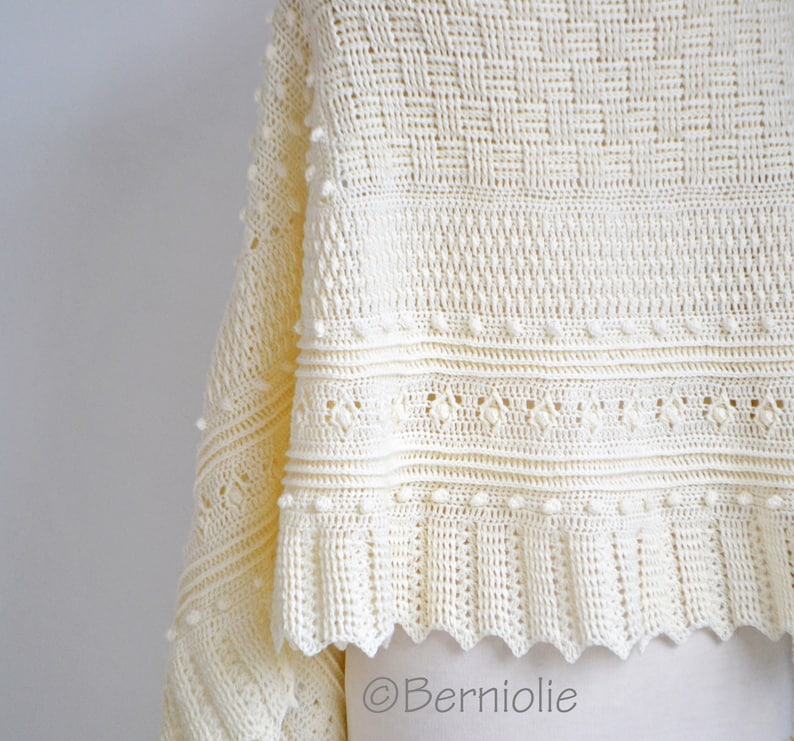 May include: A cream-colored crochet shawl featuring a complex pattern of textured bobbles, diamond-shaped openwork, and a ribbed border. The fabric displays a variety of intricate stitch details and a scalloped edge. The text Berniolie is visible.