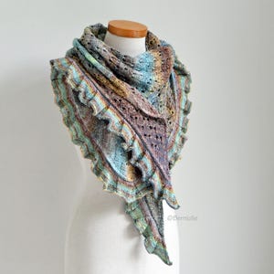 Può includere: Uno scialle lavorato a maglia multicolore con bordo a balze. Lo scialle presenta un mix di blu, verdi, marroni e viola. Lo scialle è esposto su un manichino. Lo scialle ha una forma triangolare e un delicato design a maglia traforata.