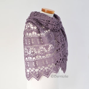 May include: A lavender crocheted shawl draped over a mannequin. The shawl features a floral pattern and a scalloped edge. The openwork design is visible throughout the shawl. The shawl is made of a soft, lightweight yarn.