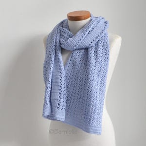May include: A light blue crocheted scarf displayed on a mannequin. The scarf has an open, lacy design and is draped to show its length and texture. The color is a soft, pastel blue, and the craftsmanship is evident in the detailed stitch work.