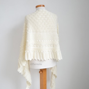 May include: An off-white crochet shawl featuring a complex pattern of textured stitches, including bobbles and a scalloped edge. The garment is displayed on a mannequin and includes the text Berniolie printed on the lower section.