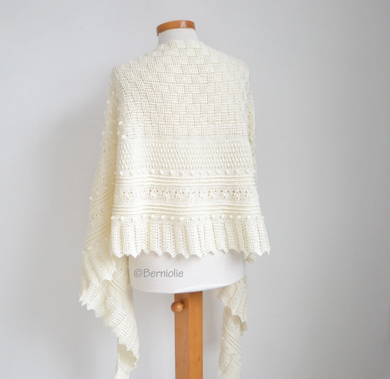 May include: An off-white crochet shawl featuring a complex pattern of textured stitches, including bobbles and a scalloped edge. The garment is displayed on a mannequin and includes the text Berniolie printed on the lower section.